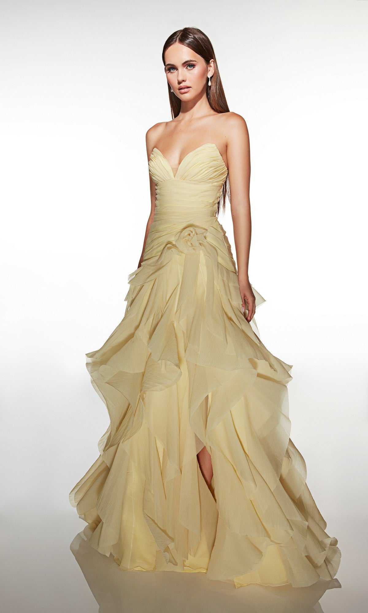 A woman stands against a plain background wearing the Alyce 62024, a strapless, floor-length pale yellow prom dress with a ruffled layered skirt.